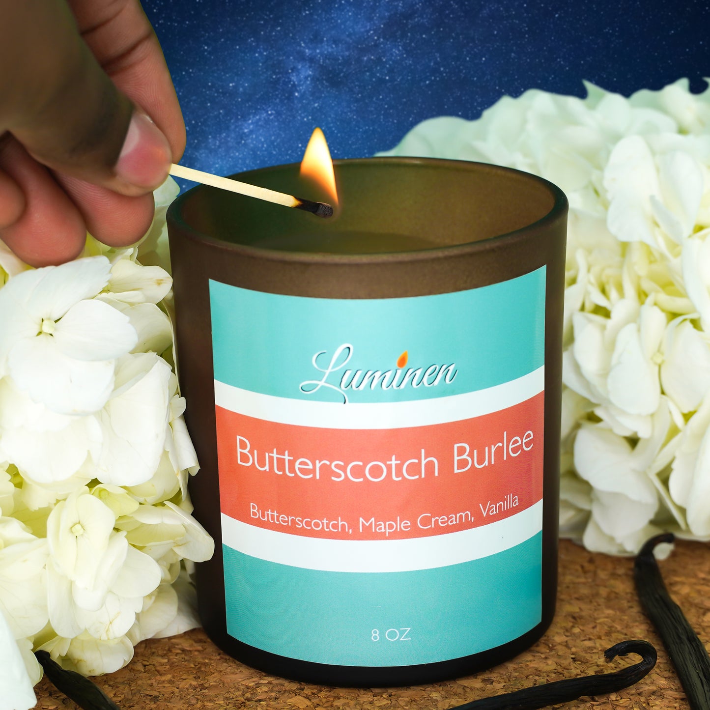 A brown candle named Butterscotch Burlee with a star filled background, flowers and vanilla sticks being lit with a match