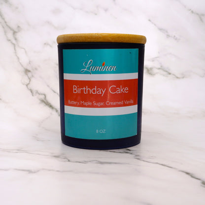 A Birthday Cake candle in a dark blue frosted  jar with a bamboo lid against a marble background